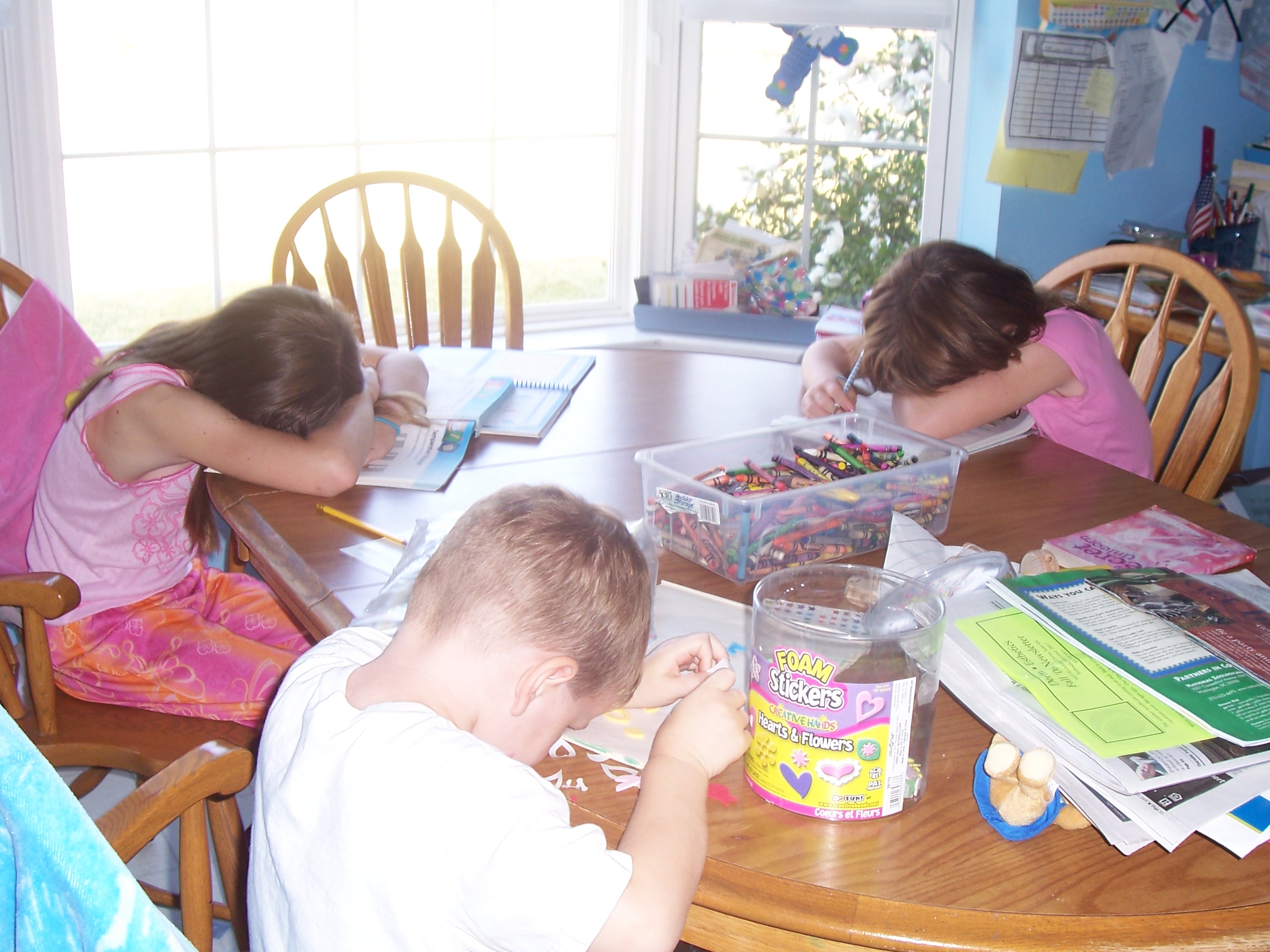 Kids Work on Summer Workbooks at the Kitchen Table. All three are resting their heads on their hands. Their hands are on top of their workbooks.